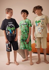 Trois garçons portent des vêtements à thème Minecraft : un t-shirt noir avec un short, un t-shirt vert avec un short imprimé, et un ensemble t-shirt beige avec short.