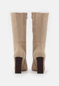 Beige läder knähöga stövlar med en strukturerad design, guld dragkedjor och en staplad träklack. Texturerad panelering på baksidan.