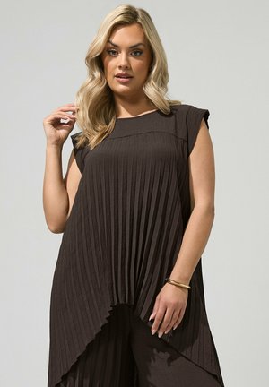 Femme aux cheveux blonds portant une tunique plissée sans manches sombre et un pantalon assorti, posant sur un fond clair uni.