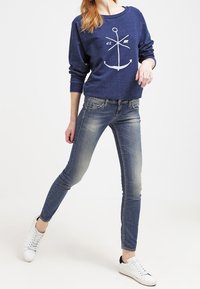 Sudadera azul marino con gráfico de ancla blanca, combinada con jeans ajustados azul desgastados y zapatillas blancas con detalles negros.