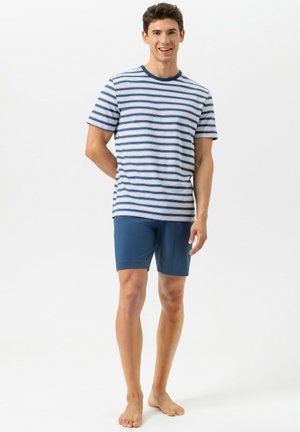 Jonge man staat blootsvoets, draagt een gestreept shirt met korte mouwen en blauwe korte broek, glimlacht met één hand achter zijn rug.