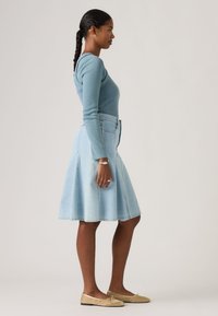 Γυναίκα σε πλαϊνή όψη που φοράει μπλε μακρυμάνικη μπλούζα, ανοιχτόχρωμη τζιν φούστα μέχρι το γόνατο, χρυσά επίπεδα παπούτσια και πλεξούδες στα μαλλιά.