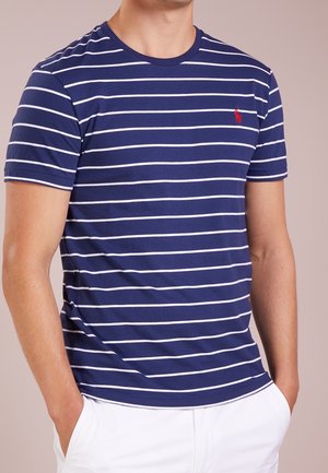Uomo che indossa una maglietta a maniche corte blu navy con strisce orizzontali bianche e piccolo logo rosso, abbinata a pantaloni bianchi, con le mani nelle tasche.