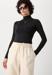 Čierny dlhý rukáv turtleneck top spárovaný s béžovými elastickými nohavicami. Materiál je hladký; výstrih má zakrivený dizajn.