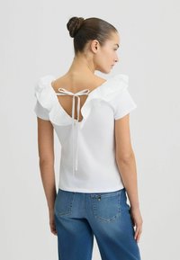 T-shirt bianco in cotone con spalle arricciate e schiena a chiave, legato con un nastro bianco, abbinato a jeans in denim blu.