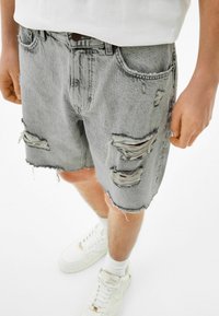 Henkilö, jolla on päällään vaaleanharmaat kulutetut farkkushortsit, joissa on repsottavat reunat, sekä valkoinen paita. Asusteena myös valkoiset tennarit ja sukat.