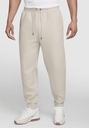 Homme portant un pantalon de survêtement beige avec une taille élastique et un cordon, associé à des baskets blanches de sport.
