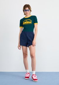 Green short-sleeve shirt with yellow accents and text, paired with navy striped shorts. Model wears red sneakers with pink laces.