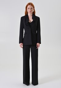 Completo nero su misura caratterizzato da un blazer con collo a cowl, chiusura a un bottone e due tasche frontali, abbinato a pantaloni a gamba larga.