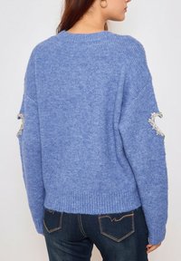 Pull en maille bleu clair à coupe décontractée, avec un ourlet côtelé et des accents décoratifs en argent sur les manches. Texture douce, design décontracté.