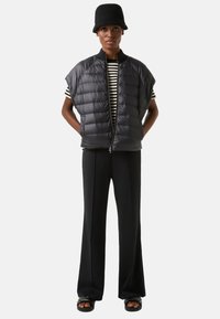 Czarna pikowana kamizelka z dużymi otworami na ręce, zestawiona z pasiastą bluzką, czarnymi spodniami i czarnym kapeluszem typu bucket. Stylizację dopełniają płaskie sandały.