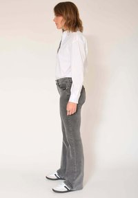 Femme debout de profil portant une chemise blanche boutonnée, un jean gris évasé et des baskets blanches à rayures noires, sur un fond uni.