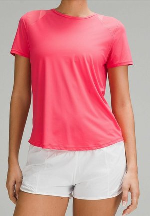 Osoba w koralowo-różowej sportowej koszulce z krótkim rękawem i białych szortach stojąca na tle jednolitego jasnoszarego tła.