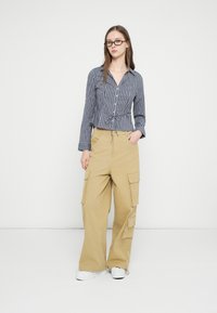 Νέα γυναίκα με γυαλιά, πουκάμισο με καρό σχέδιο και μακριά μανίκια, μπεζ παντελόνι cargo και λευκά αθλητικά παπούτσια, στέκεται με το ένα χέρι στην τσέπη.