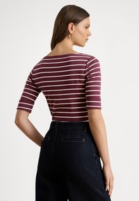 Žena s hnedými vlasmi, oblečená v pruhovanom tričku bordovej a bielej farby s rukávmi do lakťov a vo vysokých tmavých džínsových nohaviciach, otočená doprava.