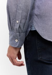 Koszula z niebieskiego chambray z kołnierzykiem zapinanym na guziki, złożonym mankietem, subtelną fakturą i zaokrąglonym dołem, w połączeniu z ciemnymi dżinsami.