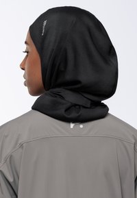 Profilo laterale di una persona che indossa un hijab Kamak nero e una giacca grigia con un piccolo logo bianco sul colletto posteriore.
