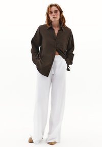 Camicia marrone con bottoni e tasca abbinata a pantaloni bianchi a gamba larga. L'outfit presenta una vestibilità comoda e un tessuto testurizzato, completato da scarpe beige.