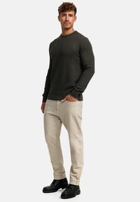 Dunkelgrüner strukturierter Strickpullover, beige Hose, schwarze Stiefeletten. Der Pullover hat ein Zickzackmuster und gerippte Bündchen.