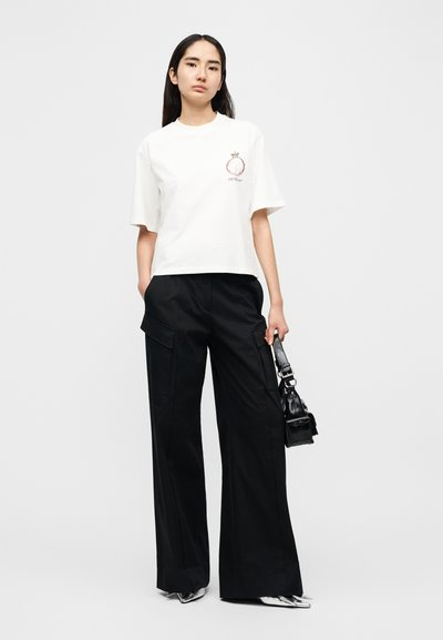 Λευκό κοντό t-shirt με μεταλλικό γραφικό και κείμενο, σε συνδυασμό με φαρδιά μαύρη cargo παντελόνια και μικρή μαύρη τσάντα χειρός.