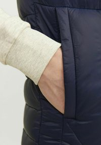 Mornarska pufasta jakna s prišitim vzorcem, ki ima stransko žep. Rokav ima lahek rebrast manšeti, kar dodaja kontrast teksturi.