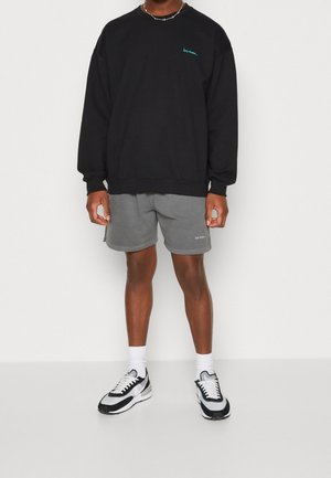 Homme portant un sweat-shirt à manches longues noir avec un petit texte bleu, un short gris avec un texte blanc, des chaussettes blanches et des baskets noir et blanc.