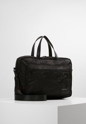 Serviette Diesel noire à motif camouflage avec double poignée, poche zippée à l'avant et bandoulière détachable, posée sur un piédestal blanc.