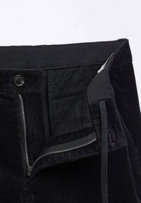 Pantalon en velours côtelé noir avec une fermeture éclair ouverte et un bouton, montrant le tissu intérieur et un cordon noir à la taille.