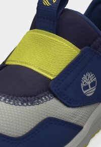 Gros plan sur une basket bleue, grise et jaune avec une bride auto-agrippante et un logo d'arbre sur le côté.