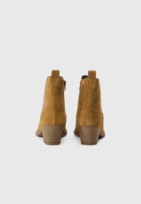 Botas de tornozelo em camurça castanha com biqueira arredondada, salto curto empilhado e fecho de correr. Inclui uma aba para facilitar o uso. Superfície texturada.