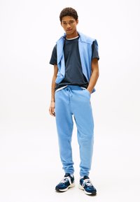 Svetlo modre trenirke s pasom na vrvico, kombinirane z mornarsko modro majico s kratkimi rokavi in svetlo modrim puloverjem, premetanim čez ramena. Črno-modri supergi.