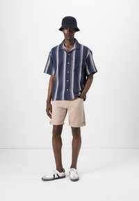 Chemise à manches courtes, à boutons, avec motifs bleu marine et rayés, associée à un short beige et des baskets blanches. Un chapeau seau noir complète le look.
