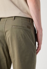 Khaki trousers with a slim fit, featuring a back pocket and subtle logo detail. The fabric has a smooth texture and minimal design.