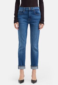 Jean en denim bleu à taille haute, jambes droites et revers retournés. Présente un léger délavage et deux poches avant.