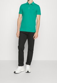 Polo vert avec un col et une patte de boutonnage à trois boutons, associé à un pantalon noir et des baskets blanches, avec un design simple.