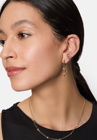 Gouden ringoorbel met een hangende ketting en kleine stenen, gecombineerd met een slanke gouden ketting met subtiele accenten, gedragen op donkere stof.