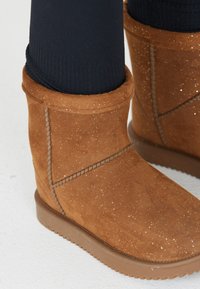 Bottes chevilles en suede marron avec des paillettes subtiles, dotées d'une texture lisse et d'un bout arrondi. Accents cousus et semelle en caoutchouc pour une meilleure adhérence.