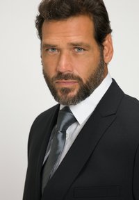 Uomo in un abito nero con camicia bianca, che indossa una cravatta grigia a fantasia. Ha una barba e capelli corti e ondulati; l'espressione è seria.