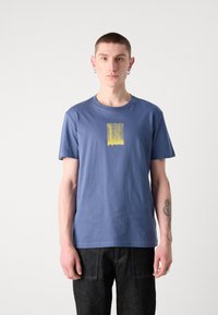 T-shirt de algodão azul com decote redondo, apresentando um design de salpicos de tinta amarela no centro. Usado com jeans escuros.