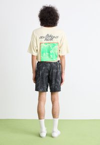 T-shirt oversize beige con una grafica verde e arancione sulla schiena, abbinata a pantaloni corti neri a fantasia e sneakers bianche con calzini alla caviglia.