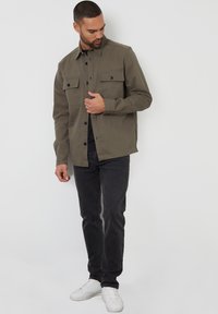 Olijf groene knoopjack met twee borstzakken, zwarte knopen en een ontspannen pasvorm, gecombineerd met donkere jeans en witte sneakers.