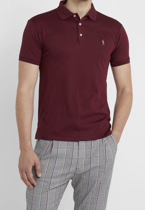 Bordeauxrotes Poloshirt mit klassischem Kragen, drei Knöpfen und kleinem gesticktem Logo. Kombiniert mit grauen karrierten Hosen mit schwarzen und roten Akzenten.
