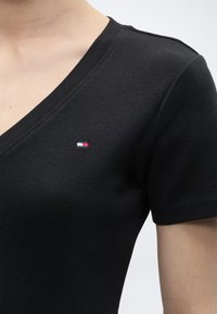 Κοντινό πλάνο ενός ατόμου που φοράει μαύρο κοντομάνικο μπλουζάκι με V λαιμόκοψη και μικρό ορθογώνιο κόκκινο, λευκό και μπλε λογότυπο στο στήθος.