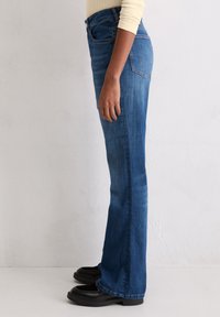 Dżinsy dzwony w kolorze niebieskim, z podwyższonym stanem, bocznymi kieszeniami i efektownym spraniem. Połączone z czarnymi butami na wsuwanie.