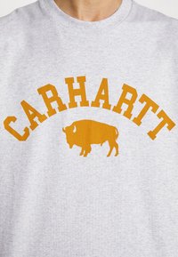 Grå sweatshirt med fedt orange "CARHARTT" tekst og en bøffel grafik nedenfor, rund halsdesign med en blød tekstur.