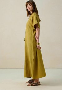 Robe maxi jaune moutarde avec une finition texturée, manches courtes larges, fente sur le côté et coupe décontractée. Associée à des sandales à imprimé léopard.