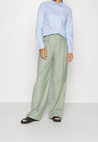 Hellblaues Hemd mit Kragen, kombiniert mit lockeren, hoch taillierten grünen Hosen. Schwarze Sandalen mit offenem Fußbett und glatte Stoffstrukturen.