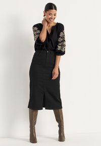 Zwarte blouse met geborduurde bloemendetails op de mouwen, gecombineerd met een zwarte midirok met een voorknos, en bruine knie hoge laarzen.