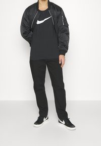 Svart bomberjacka med blank yta, ihop med en svart t-shirt med en vit Nike swoosh, svarta byxor och svarta sneakers.