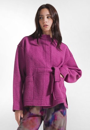 Donna che indossa una giacca di lana viola brillante con cintura e grandi tasche e pantaloni con stampa astratta colorata, guarda di lato su sfondo bianco.